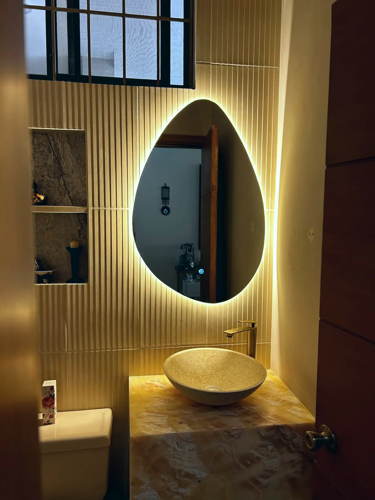 Espejo orgánico retroiluminado en un baño social, con panelado dorado y lavabo sobre encimera