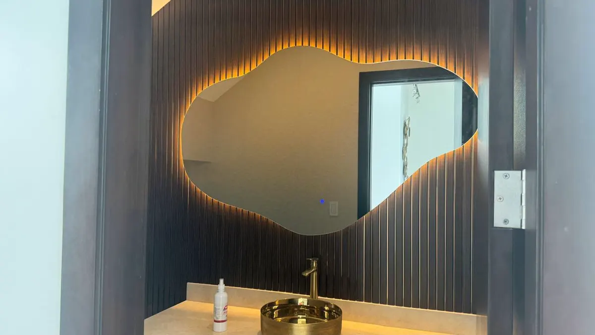 Espejo orgánico retroiluminado sobre pared azul, con grifería dorada
