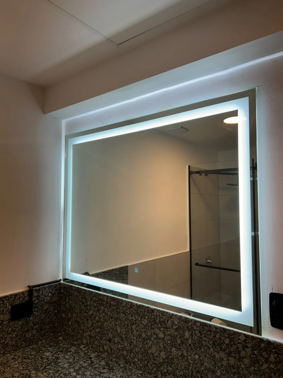 Espejo rectangular retroiluminado sobre una encimera de granito, con la mampara de la ducha reflejada
