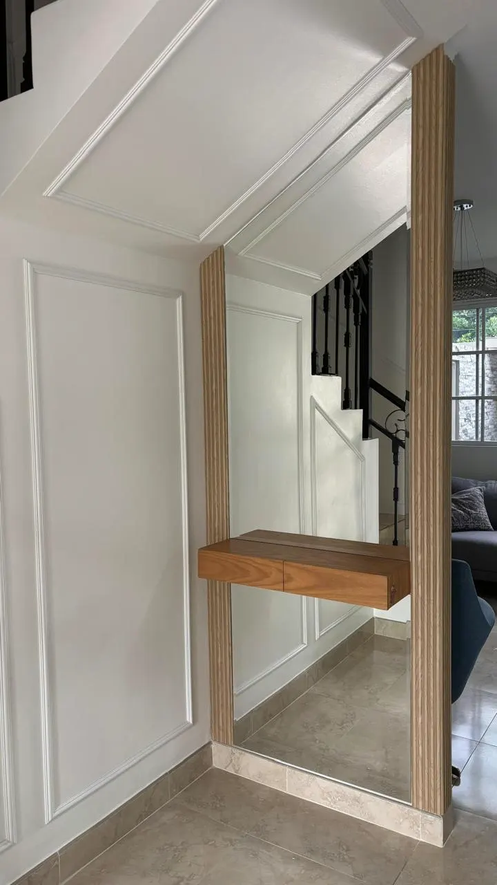 Espejo de piso a techo enmarcado en listones de madera clara, con una repisa flotante de madera, al pie de la escalera del recibidor